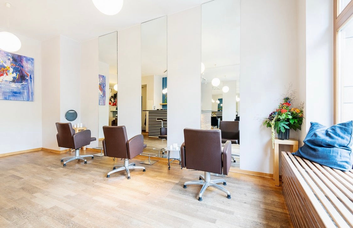 Heller, moderner Friseursalon mit braunen Stühlen, großen Spiegeln und buntem Blumenarrangement auf einer Bank.