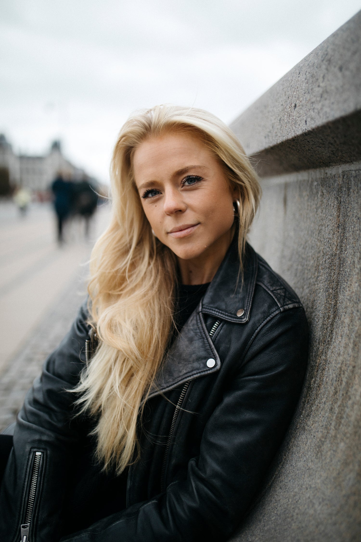 Eine Frau mit langen blonden Haaren und schwarzer Lederjacke lehnt lächelnd an einer Betonmauer.